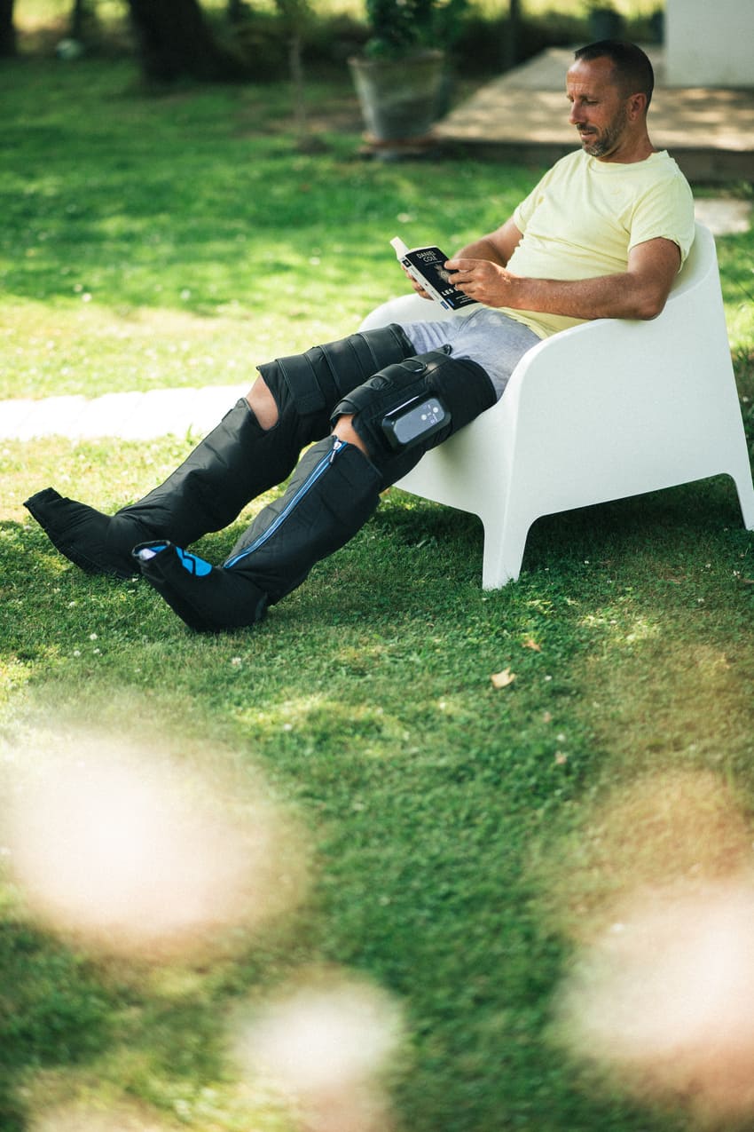 Person sitting on a white chair outdoors using a smartphone, using the Compex Ayre™ wireless compression boots on his legs