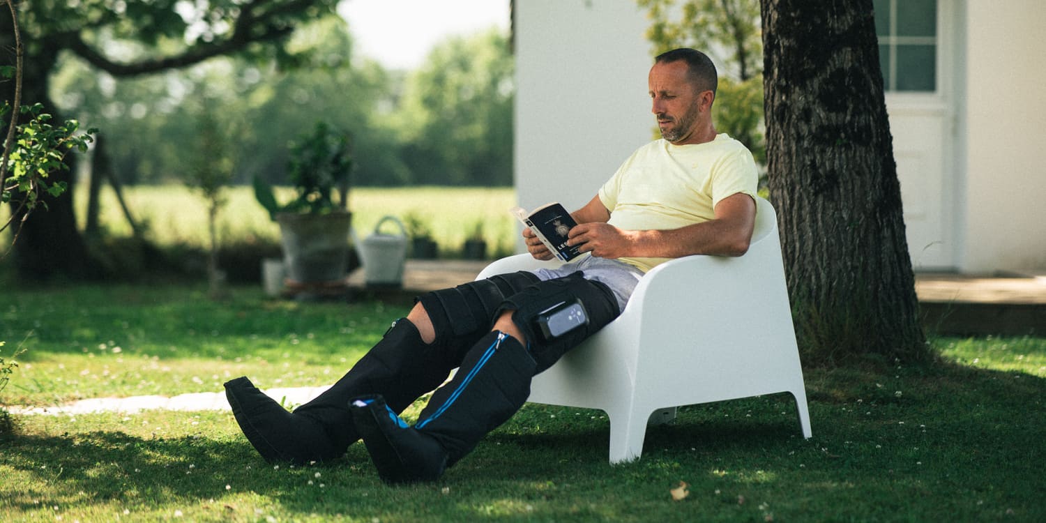 Person sitting on a white chair outdoors using a smartphone, using the Compex Ayre™ wireless compression boots on his legs