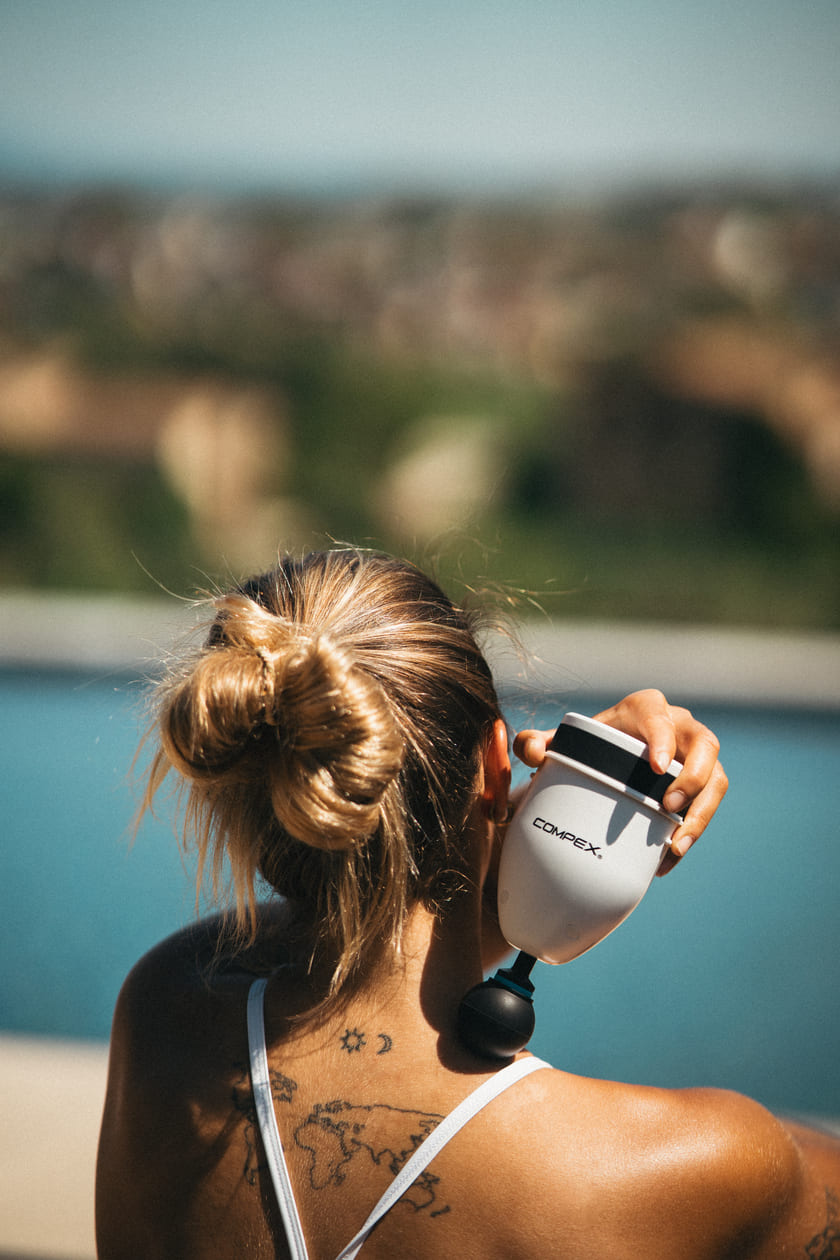 A woman using the Compex Fixx™ Mini massage gun to relax her shoulder muscles