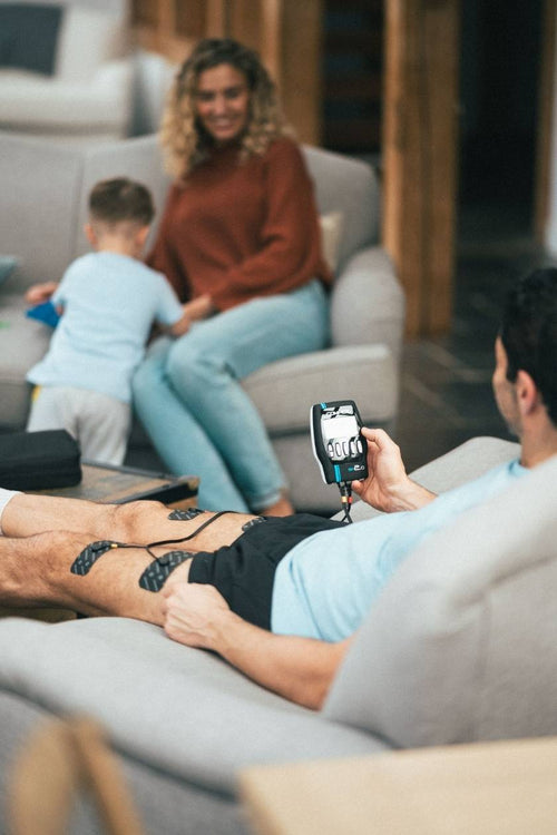 A man relaxing with his family, using the Compex SP 2.0 EMS muscle stimulator for a quad muscle recovery session