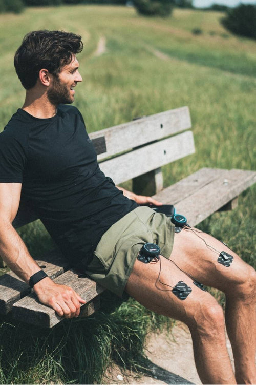 A mman sitting down outside on a bench, using the Compex Mini EMS muscle stimulator for a quad muscle recovery session