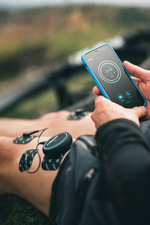 A woman sitting down outside using the Compex Mini EMS muscle stimulator for a quad muscle recovery session