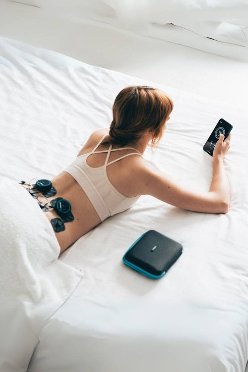 A woman lying down in bed using the Compex Mini EMS muscle stimulator for a back massage session