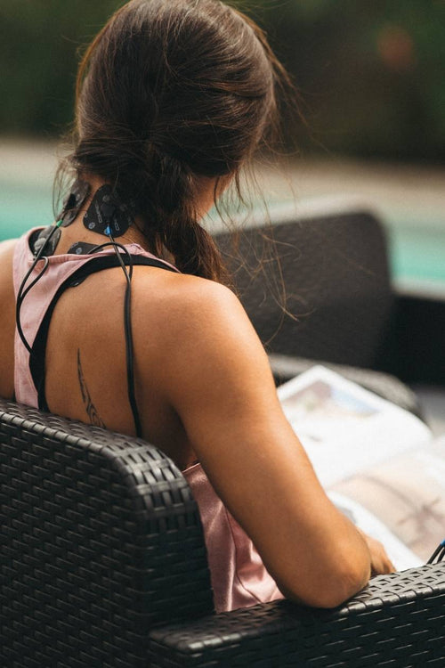 A woman sitting outside, using the Compex Sport Elite EMS muscle stimulator for a neck massage