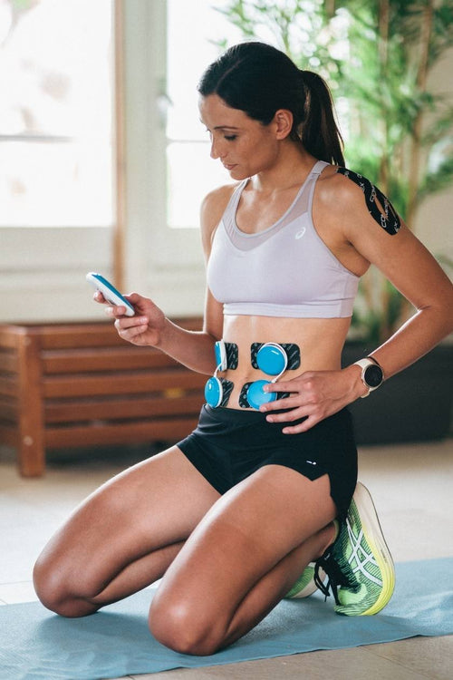 A woman kneeling, using the Compex FIT 5.0 EMS muscle stimulator to strengthen her ab muscles