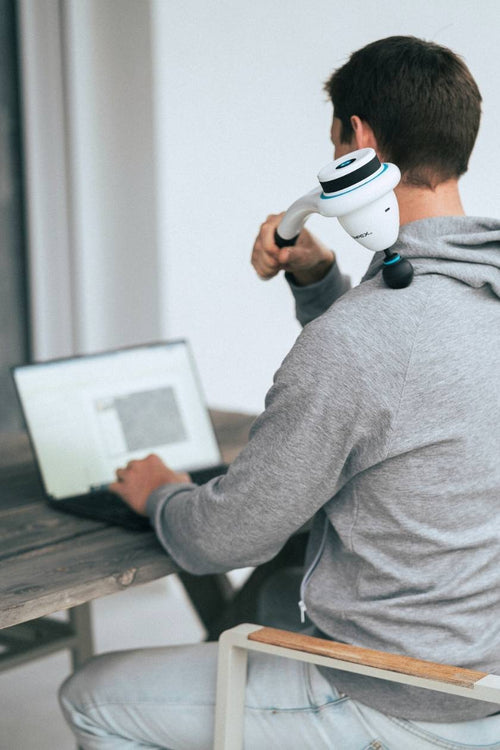 A man sitting at his laptop, using the Compex Fixx™ Mini massage gun for a shoulder massage