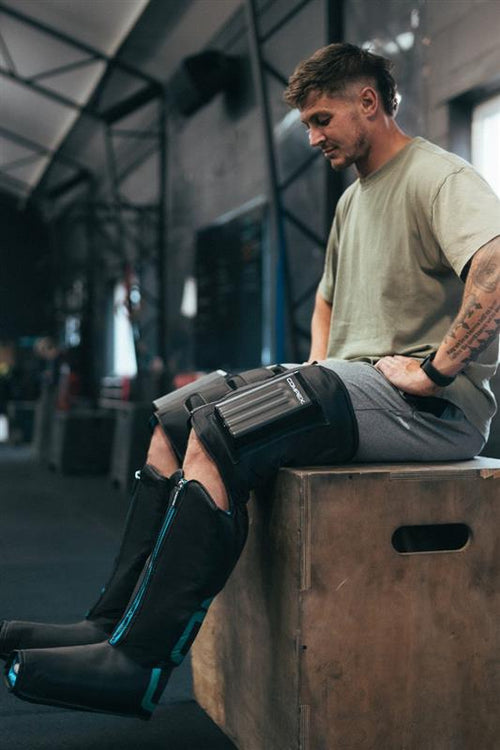 Man sitting on a wooden box wearing the Compex Ayre™ Pro wireless compression boots, in an indoor setting.
