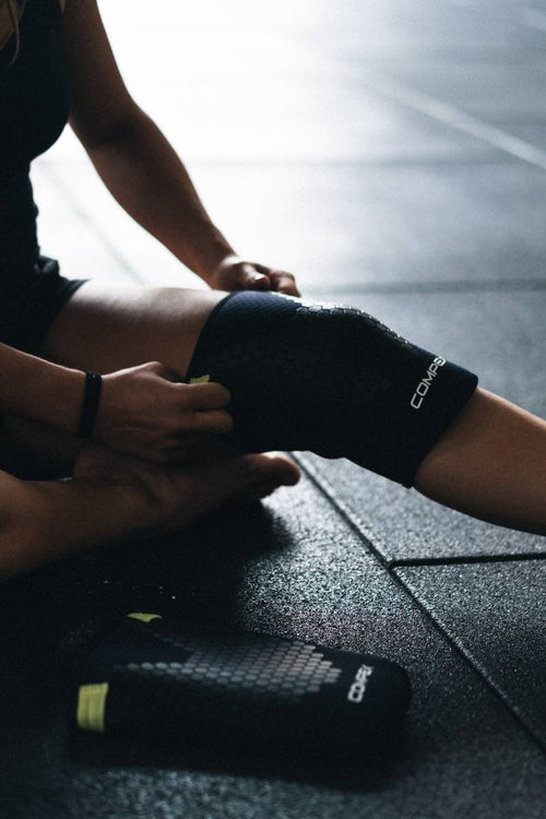 A woman sitting in a gym, putting on the Compex 5mm Knee Sleeves