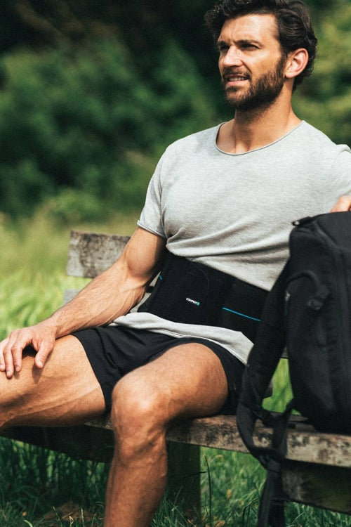 A man sitting down outside on a bench, wearing the Compex Activ'® Back Support Belt