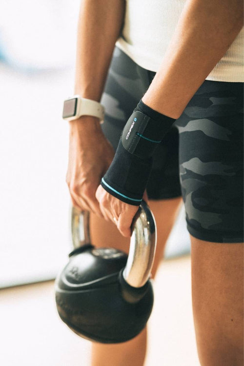 A woman lifting a kettlebell, wearing the Compex Activ'® Wrist+ Support