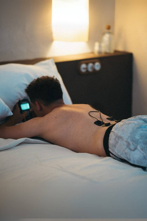 A man relaxing in bed using the Compex SP 2.0 EMS muscle stimulator to have a back massage