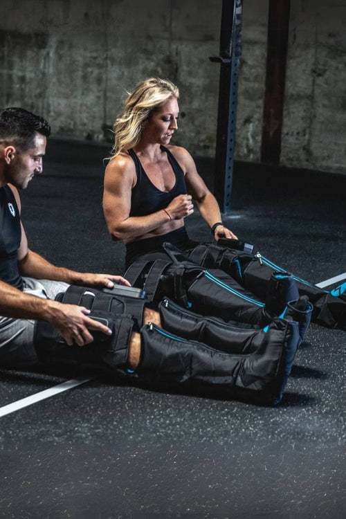 A man and woman using the Compex Ayre wireless compression boots in a gym
