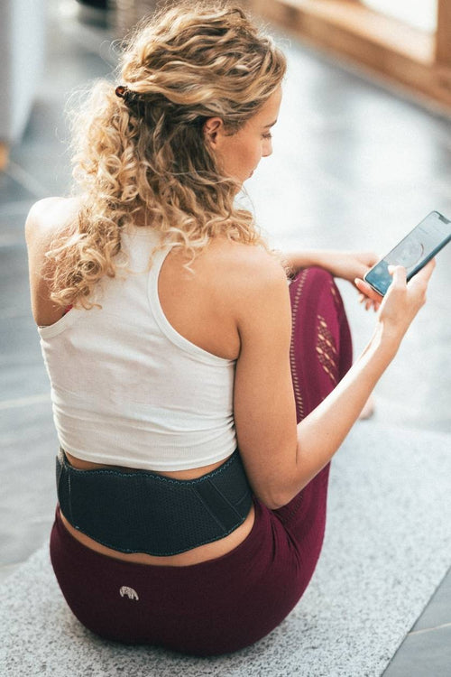 A woman sitting on a yoga mat using the Compex Corebelt EMS muscle stimulator belt