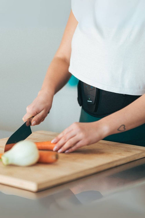 A woman chopping up vegetables whil using the Compex Corebelt EMS muscle stimulator belt to tone her abs