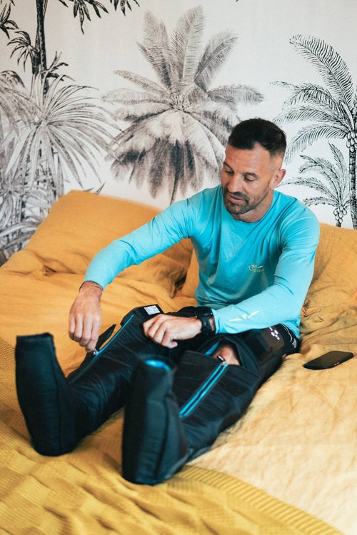 A man sitting on a bed, putting on the Compex Ayre™ wireless compression boots