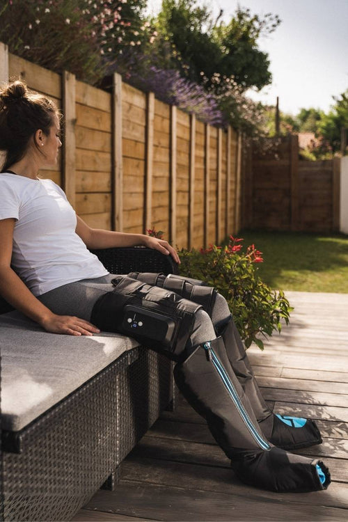 A woman sitting on a bench outside using the Compex Ayre™ wireless compression boots for leg muscle recovery