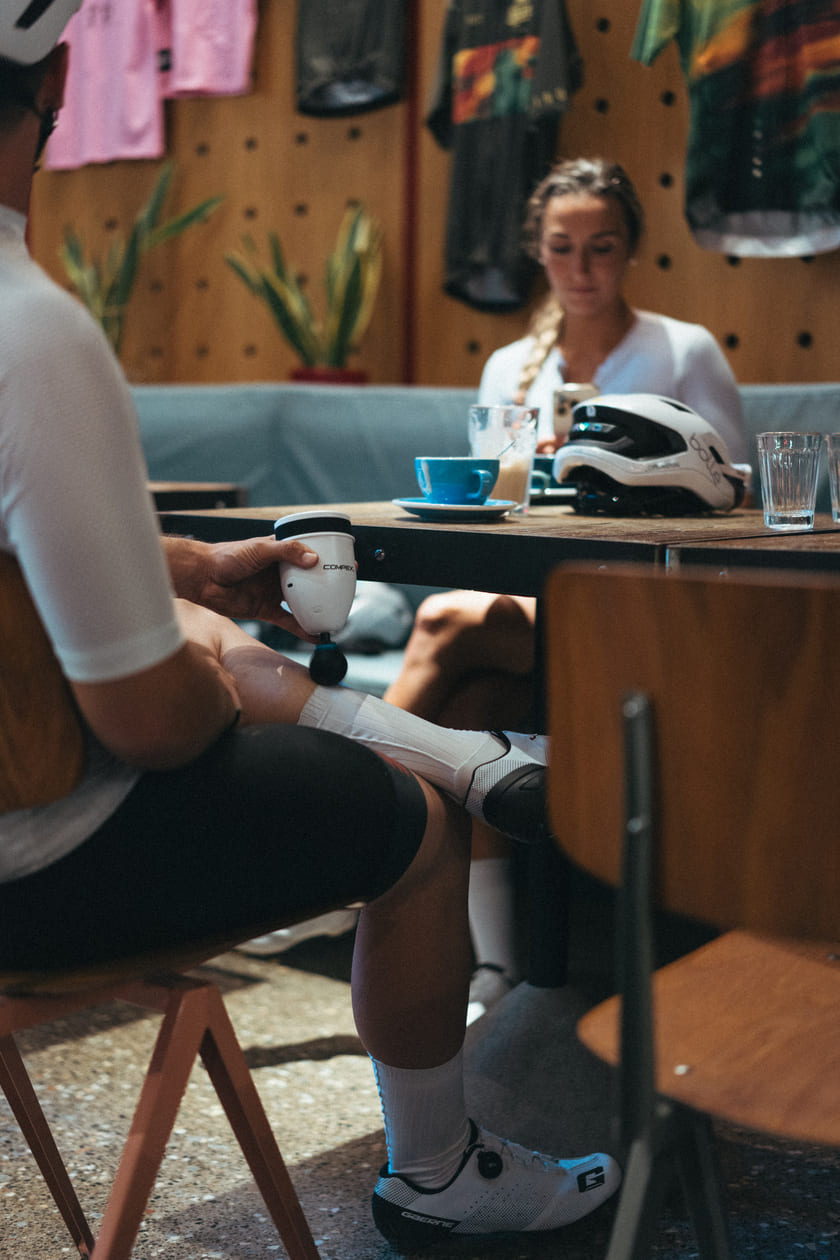 A man massaging his calf muscle in a cafe using the Compex Fixx™ Mini percussion massage gun