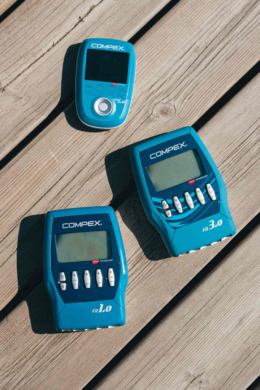 The Compex FIT 1.0, FIT 3.0 and FIT 5.0 EMS muscle stimulators on a wooden bench