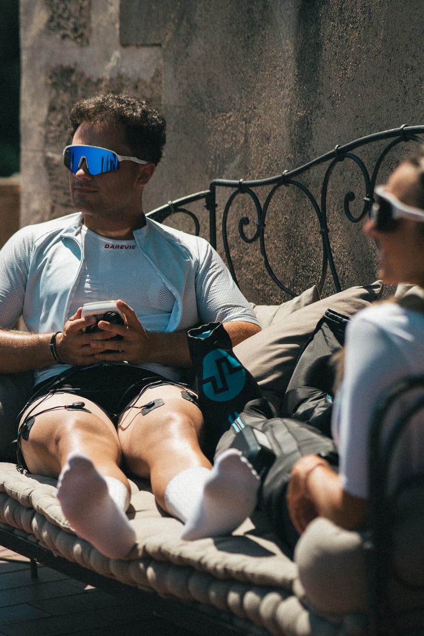 A man and a woman sitting on a bench, using the Compex SP 4.0 EMS muscle stimulator and Compex Ayre™ wireless compression boots respectively
