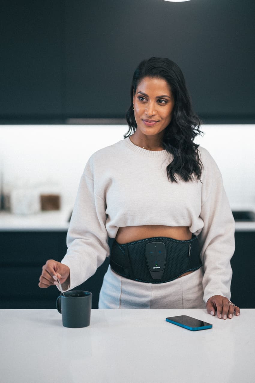 A woman standing in a kitchen using the Compex Corebelt EMS muscle stimulator belt