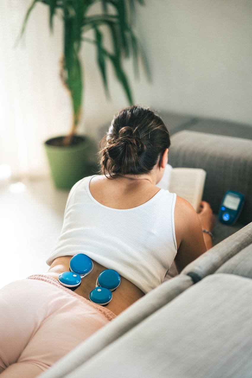 A woman relaxing on a sofa, using the Compex FIT 5.0 EMS muscle stimulator to reduce back muscle tension