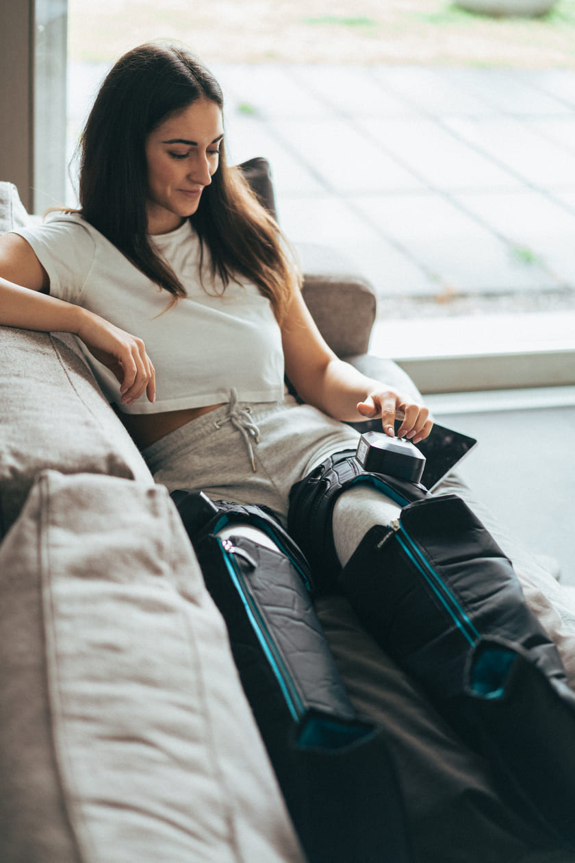 A woman sitting on a sofa using the Compex Ayre wireless compression boots